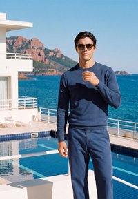 Homme portant des lunettes de soleil et une tenue bleue se tient près d'une piscine avec l'océan et des collines rocheuses en arrière-plan lors d'une journée ensoleillée.