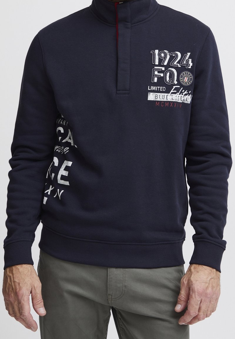 Sweat-shirt bleu marine avec col montant, ouverture zippée et accents rouges. Présente des graphismes en texte blanc et un tissu texturé. Manches longues et poignets ajustés.