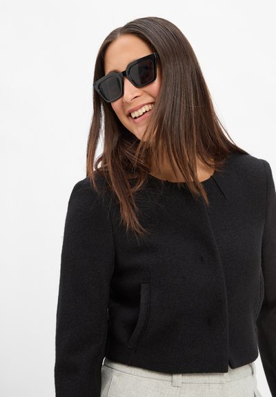 Veste noire courte avec un col rond, fabriquée en tissu texturé. Associée à des lunettes de soleil noires carrées, la personne affiche une expression légère.