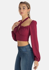 Vinröd kort blus med långa puffärmar och nyckelhålsringning, texturerat tyg, matchad med höga blå jeans.