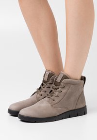 Botas de ante grises hasta el tobillo con punteras redondeadas, diseño de cordones y suelas de goma negra robustas. Costuras de acento en los bordes y lengüeta de agarre en bucle.