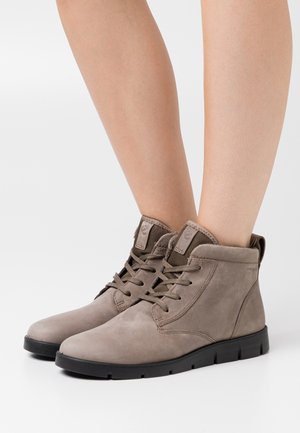 Bottines en daim gris avec bouts ronds, design à lacets et semelles en caoutchouc noir robustes. Surpiqûres sur les bords et sangle de tirage en boucle.