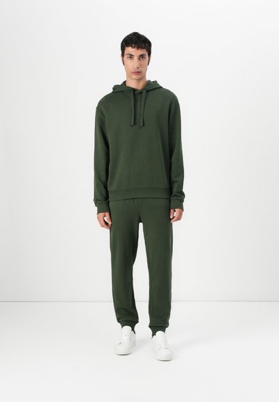 Haut sweatshirt vert foncé avec poche avant et cordon de serrage, associé à un pantalon ajusté assorti. Des baskets blanches complètent la tenue.
