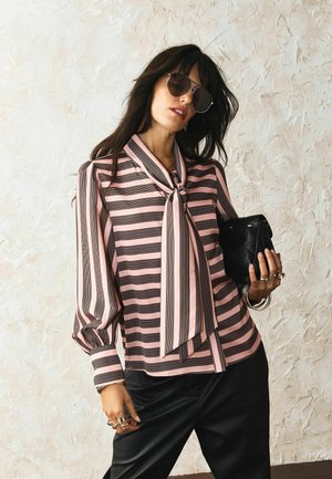 Vrouw met grote zonnebril en een roze-zwart gestreepte blouse met strik aan de hals, die een zwarte doorgestikte clutch vasthoudt.