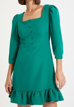 Femme portant une robe vert sarcelle avec des manches trois-quarts bouffantes, un col carré, des boutons sur le devant et un ourlet à volants, accessoirisée d'un collier chaîne en or.