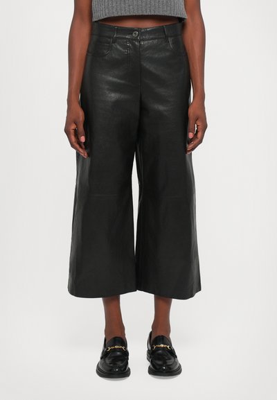 Pantalons larges en cuir noir avec une finition lisse, dotés d'une fermeture à bouton sur le devant et de poches latérales, associés à des mocassins noirs brillants.