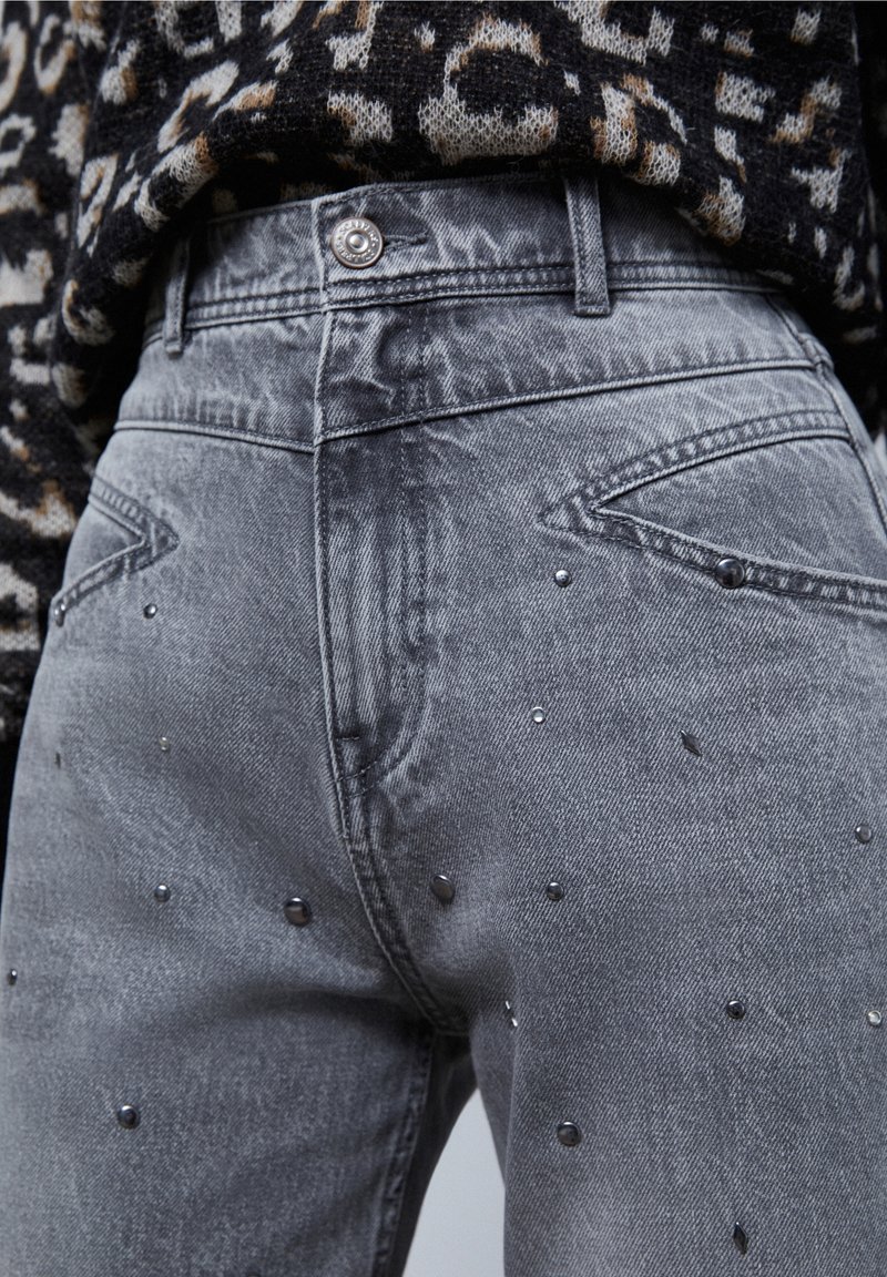 Gros plan sur un jean en denim gris avec des clous argentés et un pull noir à motif léopard rentré à la taille.