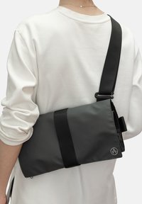 Borsa a spalla grigia realizzata in tessuto resistente, dotata di una tracolla nera regolabile, design minimale e un accento con logo sulla parte anteriore.