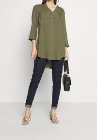 Tunique vert olive avec un col en V et des manches trois-quarts, dotée d'une patte de boutonnage, associée à un jean foncé et des escarpins à lanières. Sac à main noir.