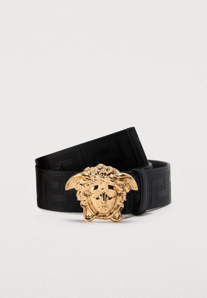 Cinturón de tela negra con superficie texturizada, con hebilla en forma de cabeza de Medusa en tono dorado y patrón geométrico a lo largo de la correa.