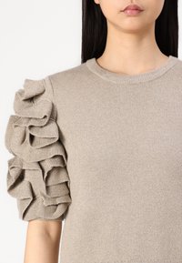Pull beige à manches courtes avec des volants superposés sur une épaule, fabriqué dans un matériau tricoté doux et texturé.