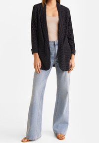 Schwarzer Blazer mit weißen Pünktchen über einem beigen Oberteil, kombiniert mit hochgeschnittenen, hellblauen Schlagjeans. Getragen mit flachen braunen Sandalen, die Arme entspannt.