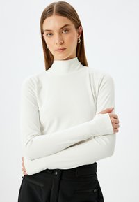 Wit, lange mouwen, hoge hals top van een gladde, rekbare stof. Model staat met de handen gekruist op de taille, draagt een zwarte broek zonder zichtbare patronen.
