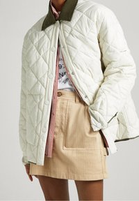 Chaqueta acolchada color crema con cuello verde, con cierres de broche y forro a rayas, llevada sobre una camisa blanca y una falda khaki.