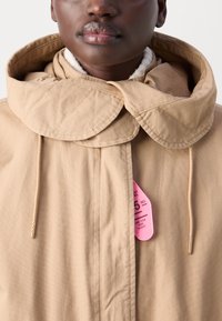 Beige quiltet jakke med en distinkt stor krage, som har hardware i gulltone og en rosa etikett festet, med rene linjer og en jevn overflate.