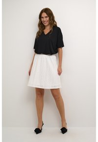 Haut noir à col en V, jupe blanche en broderie ajourée avec des motifs ondulés, et escarpins noirs à bride arrière avec des accents floraux. Texture lisse, design décontracté.