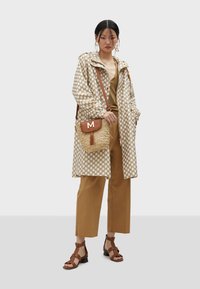 Beżowy płaszcz przeciwdeszczowy w kropki, brązowa bluzka, beżowe szerokie spodnie, brązowe sandały na paski, słomkowa torebka na ramię z beżowym klapkiem i detalem w postaci frędzli.