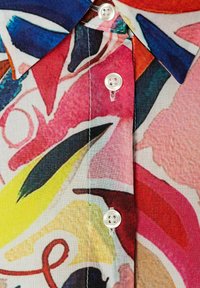 Farbige Hemdbluse mit Kragen, die ein lebhaftes abstraktes Muster in den Farbtönen Rot, Blau, Gelb und Pink zeigt, gefertigt aus leichtem Stoff.