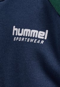 Marinblå sportklädesfabric med en texturerad yta. Har en vit "hummel"-logotyp och texten "SPORTSWEAR" på framsidan. Grön accent.