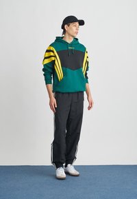 Zelená a čierna mikina s kapucňou so žltými pruhmi, doplnená čiernymi teplákmi s bielymi bočnými pruhmi a bielymi teniskami.