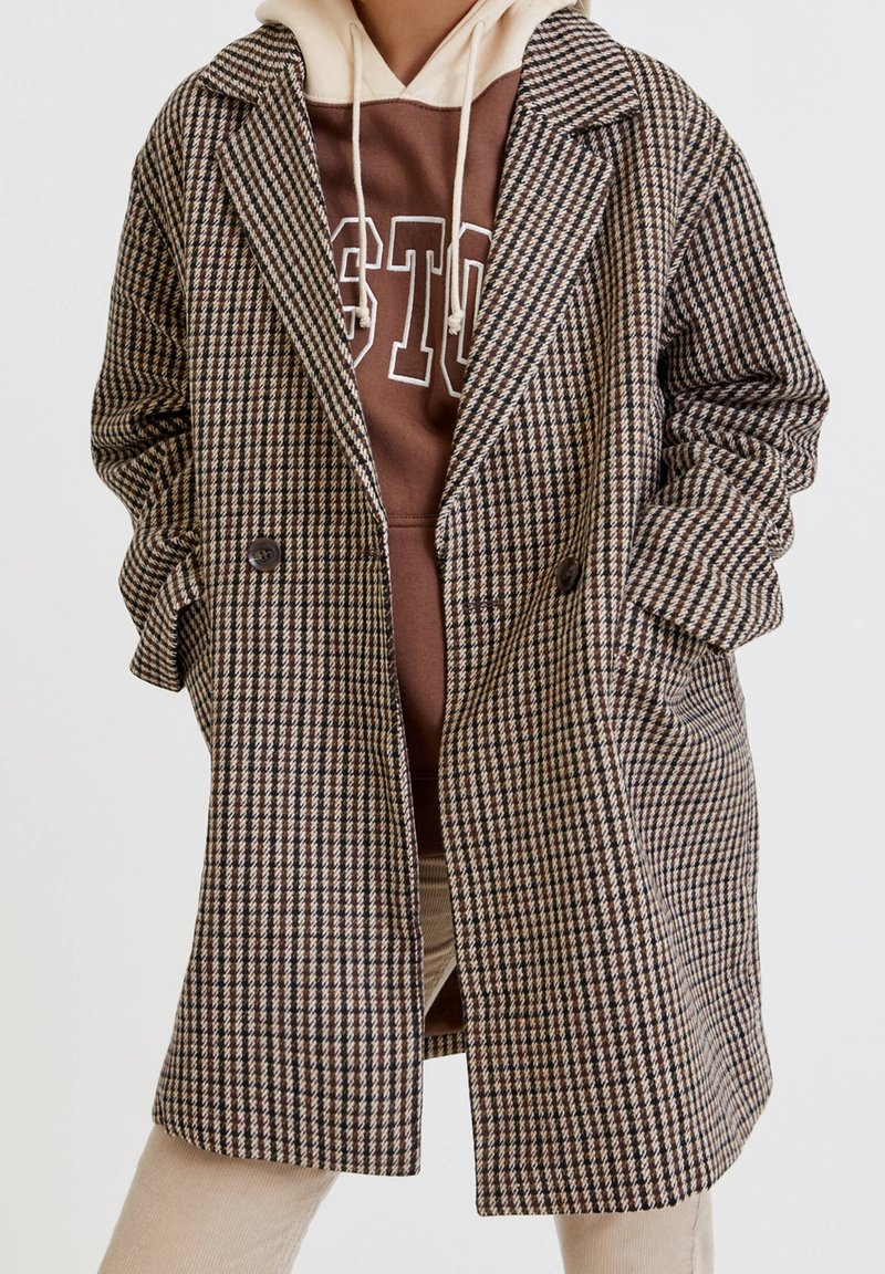 Cappotto a quadri marrone con design doppiopetto, grandi rever e tasche laterali, indossato sopra un hoodie marrone con scritte bianche.