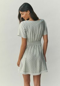 Hellgraues Kleid mit weißen Punkten, kurzen Ärmeln, geraffter Taille und ausgestelltem Rock; mit dekorativen Säumenähten. Weiche Stoffstruktur.