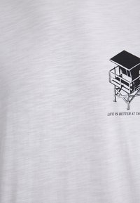 White T-shirt featuring a black graphic of a lifeguard tower and the text "LIFE IS BETTER AT THE BEACH," with a smooth fabric texture.