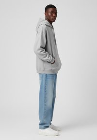 Felpa grigia con cappuccio e tasca frontale, abbinata a jeans azzurri a gamba dritta e sneakers bianche, vista di lato.