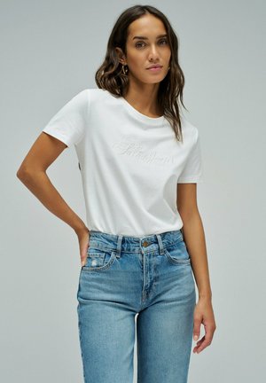 Femme aux cheveux châtains mi-longs portant des boucles d’oreilles créoles dorées, un t-shirt blanc brodé « Salsa Jeans » et un jean bleu taille haute.