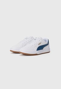 Baskets blanches avec logo Puma bleu, surface texturée, détails perforés et semelle en caoutchouc marron. Présente un design classique à lacets.