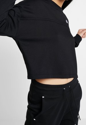 Personne portant un haut noir court à manches longues et un pantalon noir à cordon, posant avec les bras levés devant un fond uni.