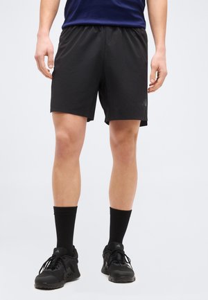 Ein Mann trägt schwarze Sportshorts, schwarze knielange Socken und schwarze Laufschuhe und steht vor einem schlichten grauen Hintergrund.