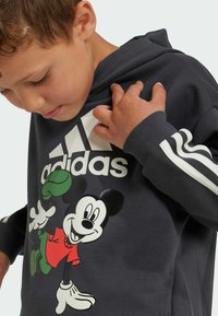 Felpa grigia con logo bianco Adidas, caratterizzata da una grafica colorata di Topolino. Include un cappuccio e dettagli a strisce sulle maniche.