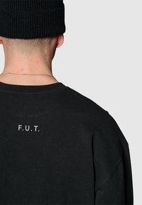 Svart bomullströja med rund halsringning och korta ärmar. Baksidan visar texten "F.U.T." i vitt. Strukturerad tyg med en avslappnad passform.