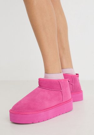 Jambes portant des chaussettes rose clair et des bottines en daim rose vif à semelles épaisses, debout sur un sol clair contre un fond uni.
