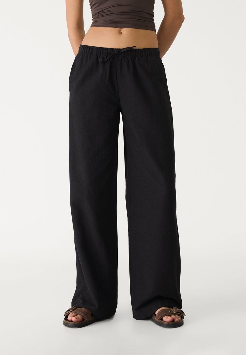 Pantalon noir, ample, avec une taille élastique et un cordon de serrage. Le tissu semble doux avec une texture lisse. Devant plat et coupe jambes larges.