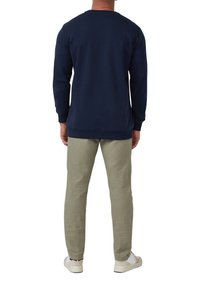 Man standing facing away wearing a navy blue long-sleeve shirt, light gray pants, and white sneakers on a white background.