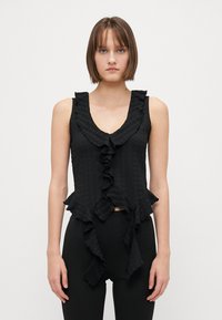 Svart ermeløs topp med volangkant i halsen og nederst. Strukturert stoff med et subtilt mønster. Tettsittende design, kombinert med svarte leggings.