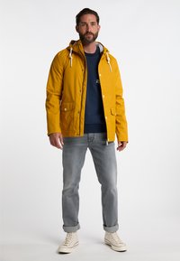 Gul vandtæt jakke med hætte, med hvide snørebånd, trykknap lommer, båret over en mørkeblå sweatshirt, parret med grå jeans.