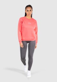 Korallfärgad sweatshirt med rund halsringning och vitt tryck, tillsammans med mörkgrå leggings och vita sneakers. Mjuk textur, avslappnad passform.