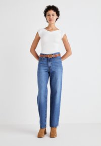 Top blanc ajusté avec des manches courtes, jeans bleus taille haute, ceinture en cuir marron avec une boucle ronde, bottes en daim marron à talon carré.