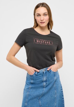 Frau trägt ein schwarzes T-Shirt mit Mustang-Logo und einen blauen Jeansrock, die Hände in den Rocktaschen, steht vor weißem Hintergrund.