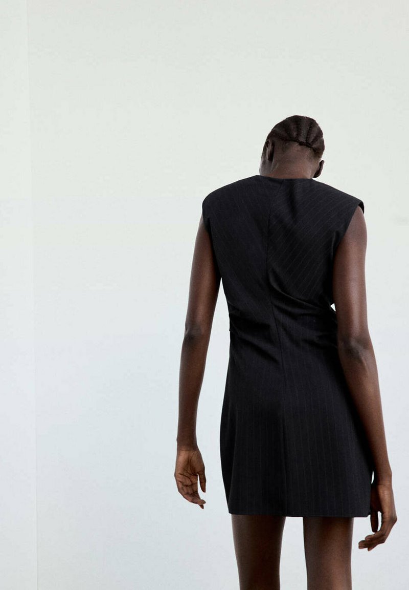 Femme aux cheveux tressés portant une robe noire sans manches, debout, dos à un mur uni de couleur claire.