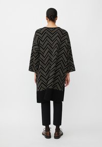 Oversized top s černým a zlatým zigzagovým vzorem, s volnými rukávy, spárovaný s černými kalhotami a tmavými, strukturovanými botami.