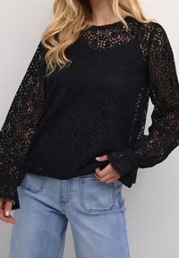 Haut en dentelle noire avec manches longues évasées, présentant un motif floral et un ourlet festonné. Associé à un jean en denim bleu clair.