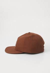 Brun stoff baseballcaps med rett skjerm, en teksturert overflate og en liten etikett foran. Sidevisning som viser den strukturerte formen.