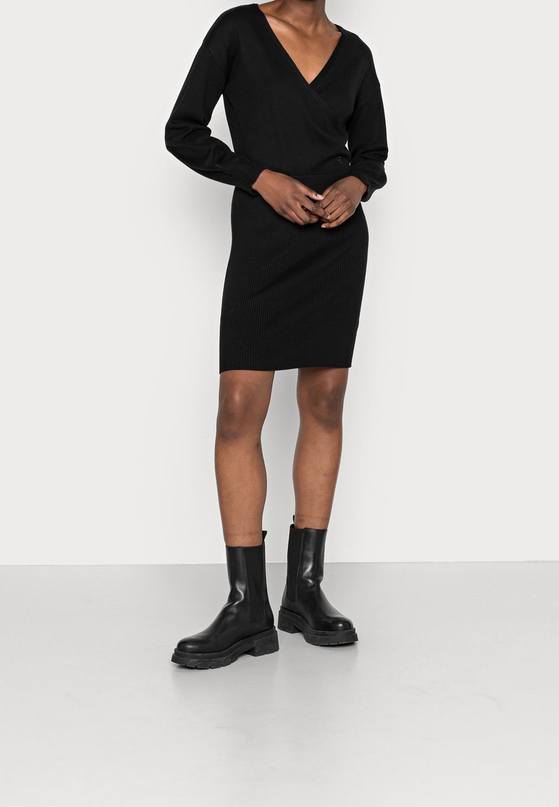 Personne portant une robe noire à manches longues et col en V ainsi que des bottines noires épaisses, debout sur un sol clair contre un mur blanc.