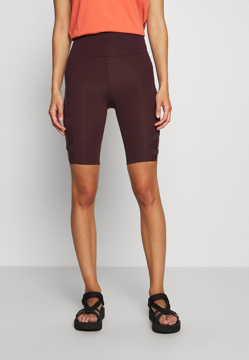 Femme portant un cuissard marron foncé taille haute et des sandales noires à plateforme, debout sur un sol gris devant un mur blanc.