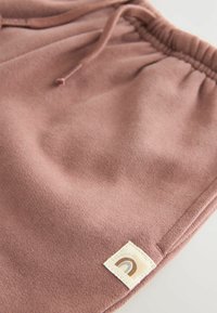Shorts en polaire rose avec une taille élastique munie d'un cordon de serrage et une étiquette cousue affichant une icône arc-en-ciel. Texture douce sur l'ensemble.