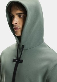 Zöld kapucnis pulóver puha anyagból, nagy kapucnival, állítható húzózsinórokkal és fekete fém kiegészítővel az elején.